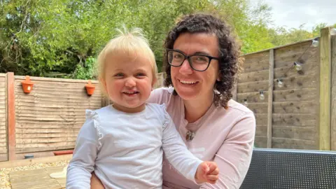 Georgie Murray-Gourlay with dark, curly hair and glasses, holding smiling baby with bright blue eyes. Sat on a bench in a garden with a fence up.