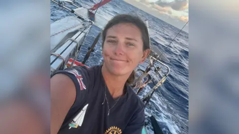 A picture showing Jasmine Harrison who is smiling and a blue t-shirt, with a Great Britain flag logo on the arm, and a yacht on the chest. She is stood on a sailing yacht which is on the water. In the background, open ocean can be seen.