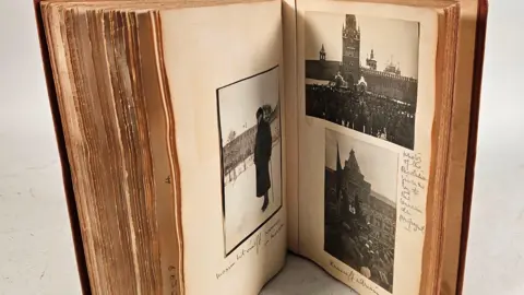 Auctioneum East Bristol & Bath Claire Sheridan's photo album standing upright on table. Three black and white photographs can be seen inside it with some handwritten notes on the pages.