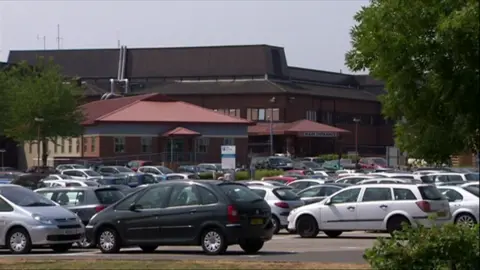 Cars parked in car park with a dark brown hospital building in the background.
