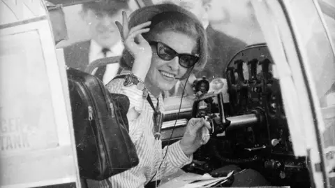 Stroud / Getty A black-and-white image of a woman with a 1960s hairstyle wearing sunglasses and sitting in the cockpit of a small aircraft. She is smiling and waving at the camera.