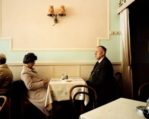 Martin Parr/Magnum Photos An elderly man and woman sitting at a restaurant table. He has a suit and combover and is smoking, she is looking down at her hands. Both are wearing 1980s clothing