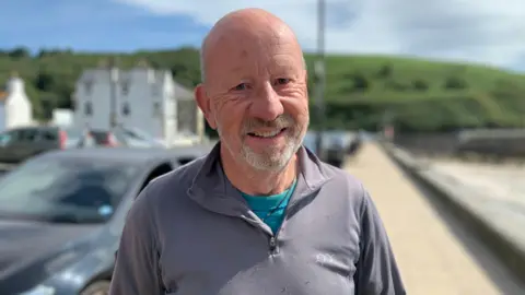 Bill has no hair and a short grey beard. He is wearing a grey quarter-zip top and is standing on the pavement of Laxey promenade with a green headline beyon on a sunny day.