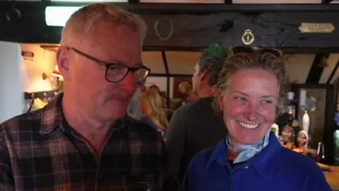 A couple called Andrew and Melissa Marsden. They are stood in a pub with people behind them. Andrew has short grey hair and black-rimmed glasses on. He is wearing a dark tartan-patterned shirt. Melissa has a broad smile on her face and fair hair swept back with sunglasses on top. She is wearing a blue jacket with a blue scarf.