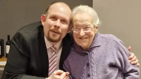 The picture shows two people standing close together indoors, with one person wearing a dark suit and a white shirt paired with a striped tie. The other person is dressed in a light purple buttoned cardigan. They appear to be holding hands in a friendly gesture, and the person in the suit has an arm around the other individual’s shoulder. In the background, there is a table with several bottles of wine placed on it against a plain grey wall.