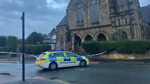 The scene in Moss Side, showing a police car and police cordon in front of a large church building
