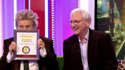 Sir Rod Stewart - a man with grey spiky hair, a black blazer, white shirt and black and white spotty tie - is holding up a certificate in a frame with his face peeking over it. Peter Davies - a man - is sitting next to him with white hair, thin rectangular glasses, a shirt and black jacket. He is smiling to the side. They are sitting on a sofa in a TV studio.