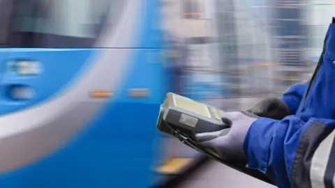 West Midlands Metro A person with a blue and black coat on and purple and black gloves is holding a ticketing machine, as a blue and silver metro blurrily speeds past in front of them.