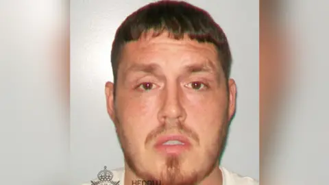 A police custody photo of Johnathan. he has short cropped brown hair and a thin reddish beard 