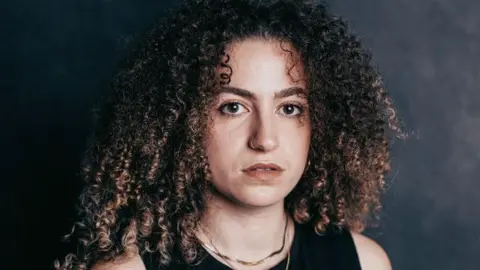Jess Douek is the lead vocalist in the Bristol band Mallavora. The image is a head and shoulders shot of her. She has curly brown hair and is wearing a sleeveless black top and a gold necklace. She is standing in front of a dark grey studio backdrop.
