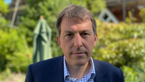 Head shot of John Glen, who is wearing a blue blazer and white and blue checked shirt. There is a green, blurred garden behind him.