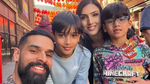 Jussi Sidhu Four family members pictured together of British Indian heritage. Dad is on the left with sleeked back black hair and a black moustache and beard. His son is next to him in the middle with straight short black hair wearing a black nike sweatshirt. His mum is to the right of him with long black hair. She is standing behind her other son who is to the far right wearing a multi-coloured vibrant Minecraft hooded sweatshirt.