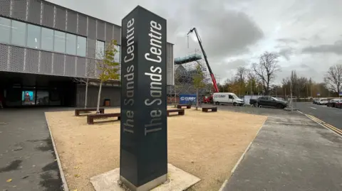 A general view of the entrance to The Sands Centre in Carlisle. A black pillar has the name of the venue written on each side in silver. The entrance to the leisure centre is visible on the left, while on the right heavy plant machinery is working on an area of the building surrounded by scaffolding.