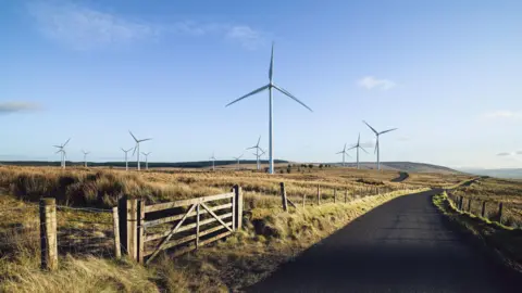 Getty Images A country road streches into the distance straddled by grass fields bordered by a wire fence. The field straight ahead has a number of wind turbines which can be seen for some distance. There is a farm gate at the front of the image leading into another field. The sky is blue and largely cloudless.