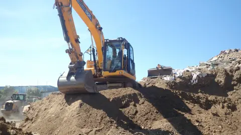 Environment Agency A mechanical digger is perched on the top of a mound of soil and waste.