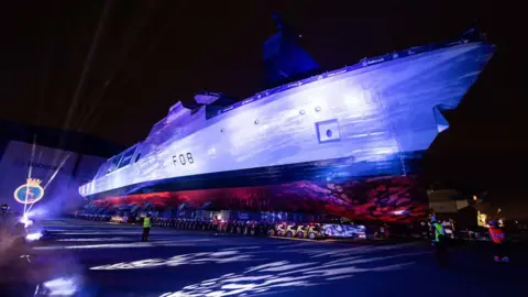 HMS Active, a large ship surrounded by strobe lighting at a dockyard with people gathered watching it.