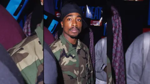 Image of Tupac Shakur wearing a blue bandana and a camouflage jacket.