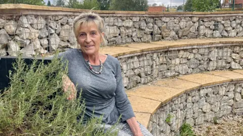 A woman with blonde hair tied back in a bun is sitting on the bottom row of a three-tiered amphitheatre next to a fern. She is wearing a stone necklace, a grey long-sleeved top and grey bottoms. The amphitheatre is made of stones secured in metal cages and topped with sand-coloured paving slabs.