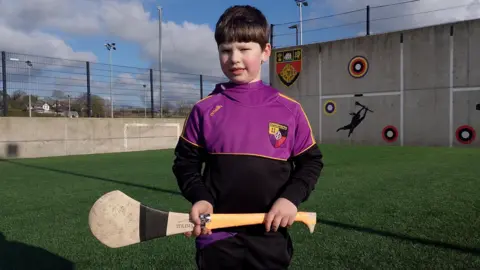 Fionn O'Neill memegang tongkat melempar di lapangan latihan. Dia memiliki rambut gelap pendek dan mengenakan pakaian olahraga hitam, ungu dan kuning dari Carryduff Gac