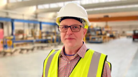 A man stands on a factory floor wearing a hardhat and glasses, as well as a hi-vis jacket. He also wears glasses. 