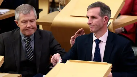 Getty Images Liam McArthur, a man with greying hair, wearing a dark suit and pink shirt, sits in the Holyrood chamber looking despondent. He is being patted on the shoulder by Willie Rennie, a man with grey hair, a grey three-piece suit and grey shirt.