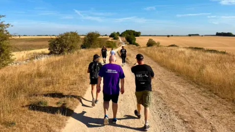 Six people with their backs to the camera as they walk along a dusty footpath that stretches into the distance with dry-looking grass and fields on both sides and a smattering of green bushes. The sky is blue with wispy clouds.