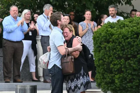 Woman cries in man's arms as people applaud in background
