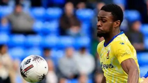 Sean Adarkwa in yellow shirt and blue shorts facing ball on the left of frame and an Oldham player in blue shirt and yellow shorts behind him