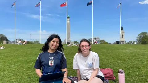 Rebecca Mugford and Veronica Estrada sat on the Plymouth Hoe Promenade. They are sat on the grass and there are five tall flags behind them, as well as a monument.