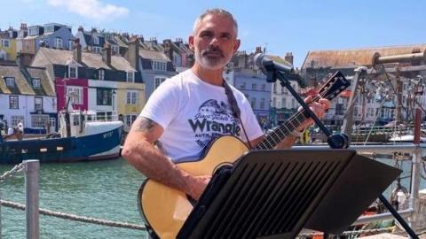 Will Adams stood with a guitar playing music by the dock, he is stood in front of a microphone and there are boats going behind him on the water