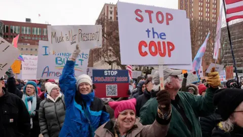 People protest against US President Donald Trump and Elon Musk outside the Michigan Capitol in Lansing, Michigan. One woman holds a sign that says: "Stop the coup". Another says: "Musk is NOT president!" The two women holding the signs are smiling.