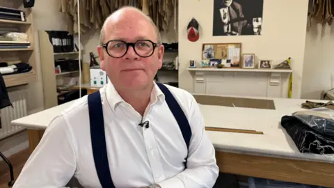 A man wearing dark glasses with a white shirt and black braces. Behind him his a tailor's shop which has material behind him and a black and white photo on the wall. 