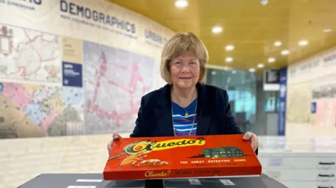 Marcia Lewis is holding one of the original versions of the Cluedo board game in its red box. She is wearing a dark blue suit jacket.