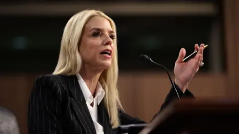 Blonde women speaks at microphone while gesturing with her left hand