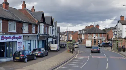 Google A general view of Derby Road in Stapleford, Nottinghamshire. Cars are parked at the side of the road next to a row of shops