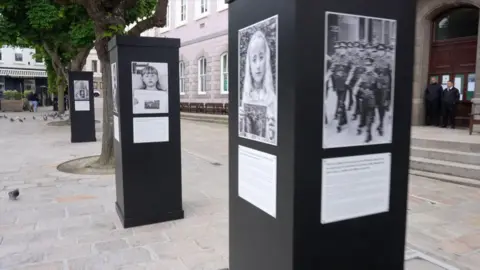 A picture of plinths in Jersey's royal square. They are black with black and white images on. They are surrounded by buildings and trees.