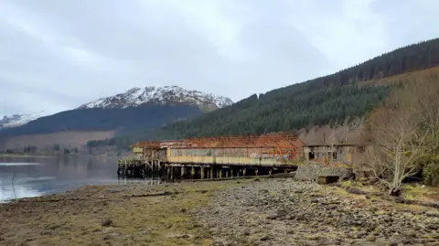 Loch Lomond & The Trossachs National Park Authority A view of the former torpedo station, sitting on the marina, with the loch visible. Rust and decay can be seen on the building even from a distance. 
