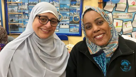 Two women wearing light coloured hijabs, they are both smiling at the camera. Behind them is  an information board with blue balloons tied to the corners