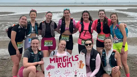 Gwen Owen A group of female runners posing in a group photo on a beach with a Môn Girls Run poster