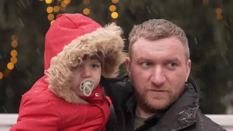 A man is holding a small child in a bright red coat with a hat, in the snow