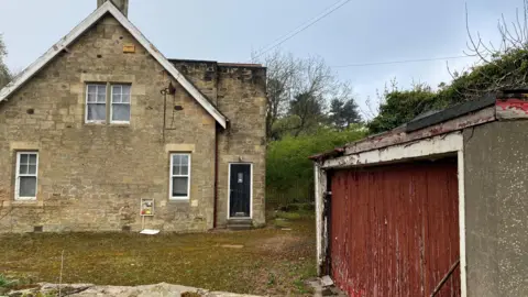 Northgate House is a large, two-storey stone built cottage of about 100 years old, but looking rather dilapidated with a drive overgrown with moss, shuttered windows and a garage with peeling paint.