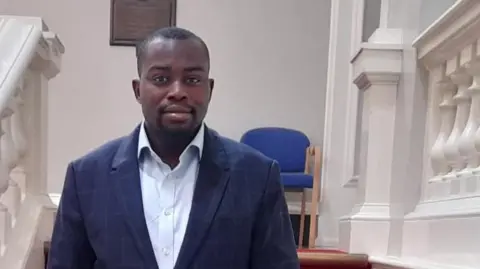 Greenwich Council Council leader Anthony Okereke wearing a suit standing at the bottom of a staircase in a council building.