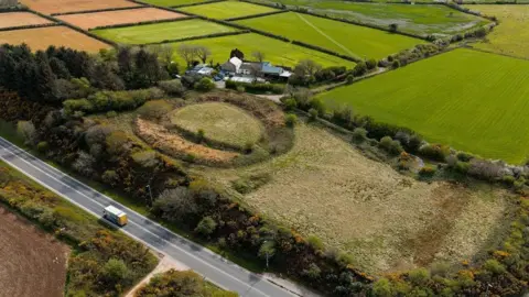 A drone shot of Castilly Henge. It is a large circle with raised ground in the middle, surrounding by trees and fields.
