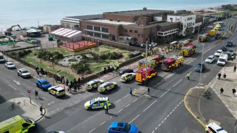 Aerial drone shot of fire engines, police cars and ambulances on a road while a fire at a brown bricked seafront building is extinguished by fire fighters.