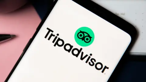 Getty Images A close up of a mobile phone with a white screen and the Tripadvisor logo on it. The logo includes a small green circle with an owl inside.
