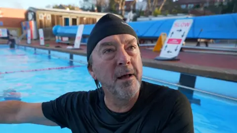 Swimmer Steve Goymer wearing a black t-shirt and a black swimming cap while standing in the pool. He is looking past the camera. 