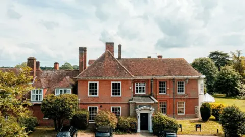 The main Stoke College building, made of red bricks with multiple chimneys. There are three vehicles parked in front of the building. Trees surround the building. 