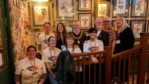 A group of Thame Pride organisers posing for a picture in front of a wall with flowery wall paper and colourful paintings