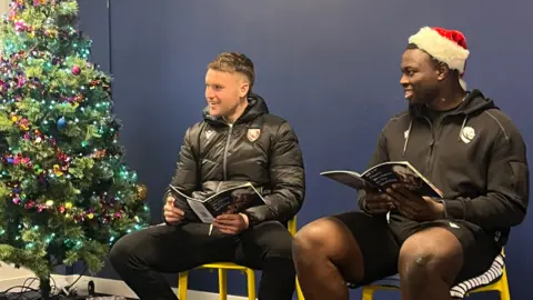 BiGDUG Two Gloucester Rugby players wearing official merchandise, with one wearing a Father Christmas hat, sit on yellow chairs against a navy painted wall. They are smiling and holding children's books as they read them. There is a decorated Christmas tree next to them.