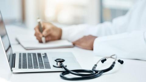 Stock photo of a laptop and stethoscope, with a doctor writing in a notebook in the background